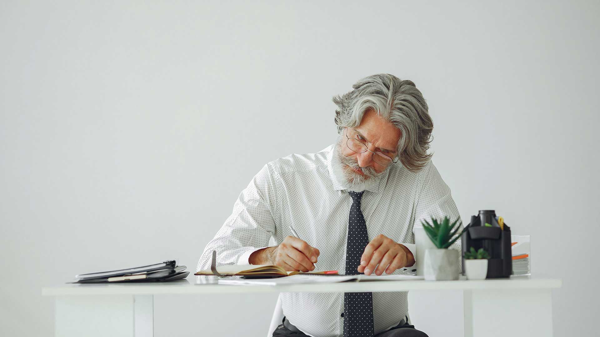 An older white man with thin rimmed glasses and medium-length grey hair is sitting at a desk and writing intently in a notebook. He is wearing a white long sleeved work shirt and navy blue tie. On the desk is a small pile of notebooks and a couple of small desk top sized plants.