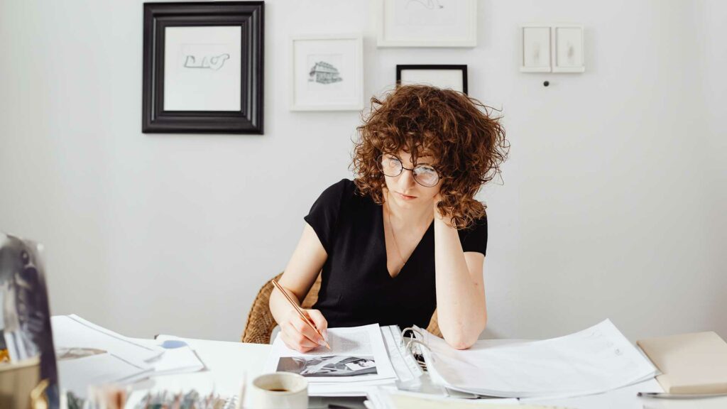 A white woman with shoulder length, curly brown hair and glasses is reading a document which looks like a research white paper. She is dressed in a black v-neck shirt and is sitting at a wooden desk writing something with a gold pen on the document in front of her. She is leaning on the document with her left elbow. Surrounding her are office supplies including notebooks and a cup of coffee.