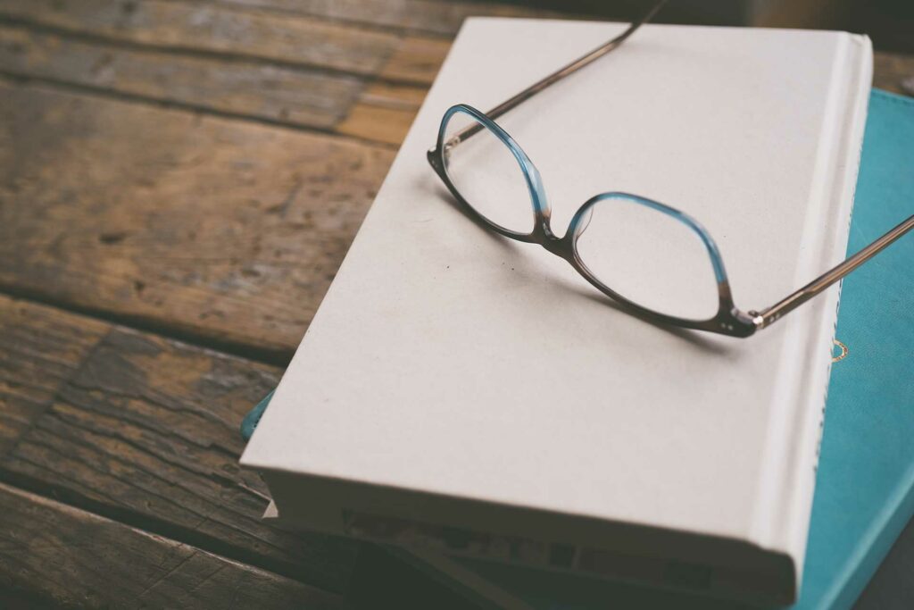 A pair of glasses rests on top of a stack of books suggesting a pause in a study session. The books are on a rough wooden surface. The book on top of the pile has a hard back white cover.