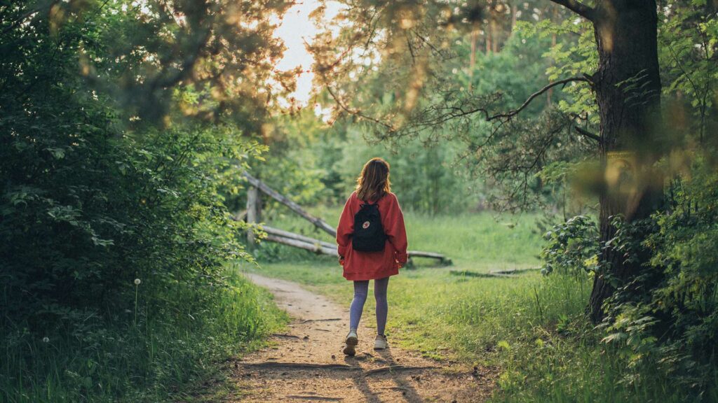 A young woman is taking part in a mindful walking exercise by walking down a country lane. She is wearing grey leggings, trainers and a red jacket with a black rucksack on her back. Sunlight is streaming down on her from between the trees above her.